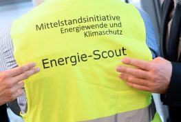 Energie-Scout in einer gelben Schutzweste. 2 gut gekleidete Herren deuten mit den Händen auf die Schrift.