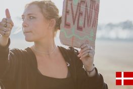 Dame mit dem Tramper Daumen und Schild in der Hand. Im Hintergrund Landschaft. Unten rechts eine Dänemarkflagge.