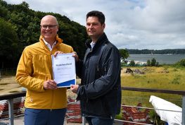 Minister Tobias Goldschmidt und Oberbürgermeister Dr. Fabian Geyer halten eine Urkunde in den Händen. Im Hintergrund befindet sich das Flächenrecycling Grundstück.