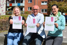 1. Flensburger Nachhaltigkeitsmarkt Vorstand, Klimaschutzmanagement und Vertreterin des FÖJ halten die Poster hoch. Im Hintergrund Häuser.